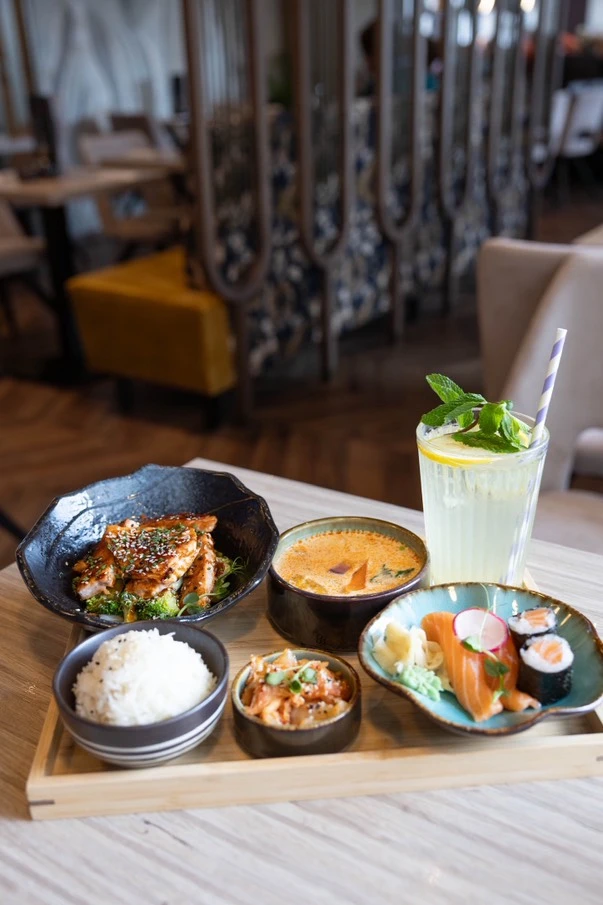 Asiatische Speiseauswahl mit glasiertem Hähnchen, Reis, Kimchi, Sushi, Sashimi und Limonade auf Holztablett.